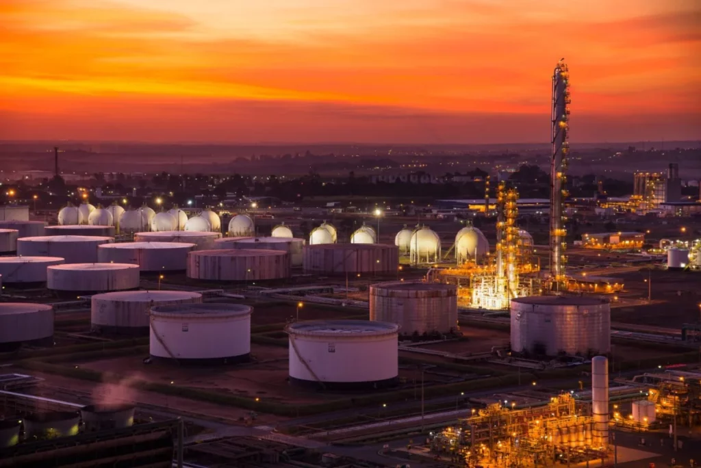 Vista aérea de refinaria no Brasil ao entardecer, com tanques e torres.