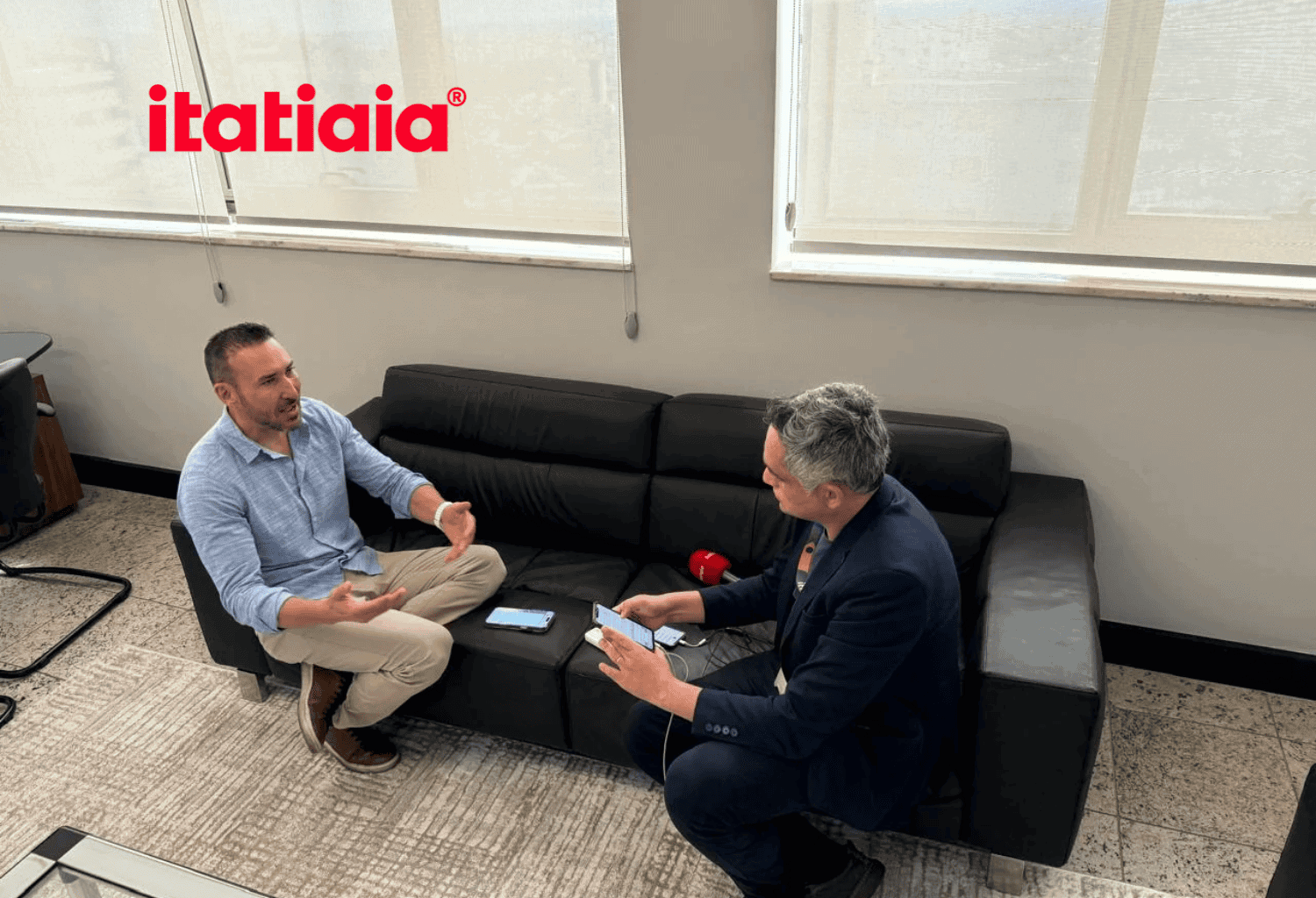 Executivo conversa com empresário em sala de reuniões, com logo itatiaia ao fundo.